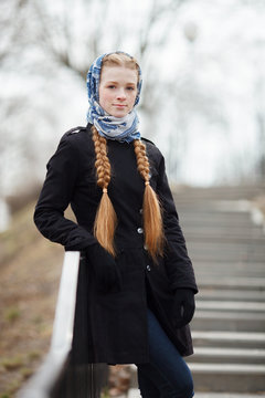 Young beautiful fashionable redhead woman with braids hairdo in blue white headcraft stylish denim black trench jacket posing on stairway in city park spring time