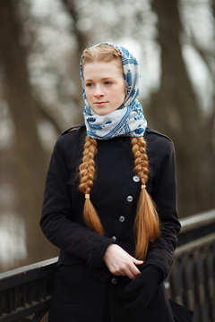 Young beautiful fashionable redhead woman with braids hairdo in blue white headcraft stylish denim black trench jacket posing on stairway in city park spring time