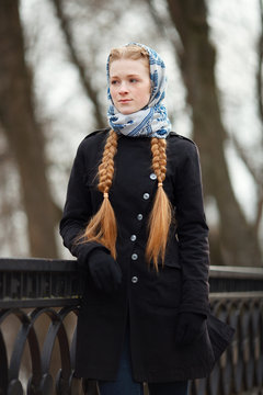 Young beautiful fashionable redhead woman with braids hairdo in blue white headcraft stylish denim black trench jacket posing on stairway in city park spring time