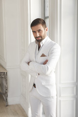 Elegant young handsome man with a beard in a white classic suit. The hotel's interior. Photo...
