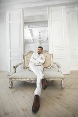 Elegant young handsome man with a beard in a white classic suit. The hotel's interior. Photo Studio. Sofa. Window. Chair. Chandelier