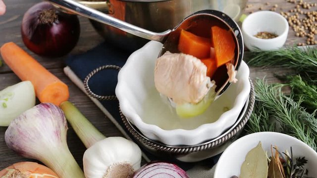 Pouring fish soup into bowl. Wooden table with roots, spices, herbs and vegetables.