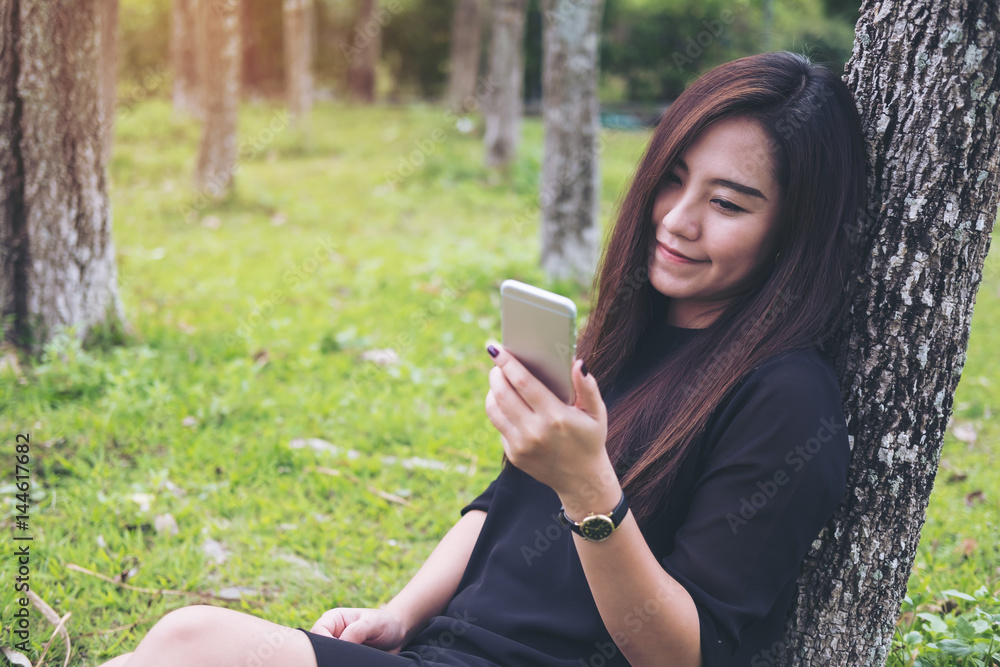 Wall mural a beautiful asian woman sitting by the tree and using smart phone with green forest background