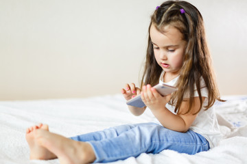 Blond kid girl playing fun with mobile smartphone on white sofa byod