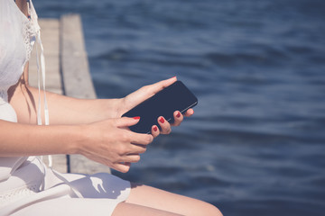 Female sitting on dock and holding blank screen smartphone. Summertime and technology concepts.