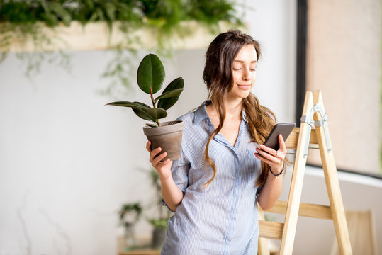 Young woman planting home with greenery standing with phone and flowerpot on the ladder