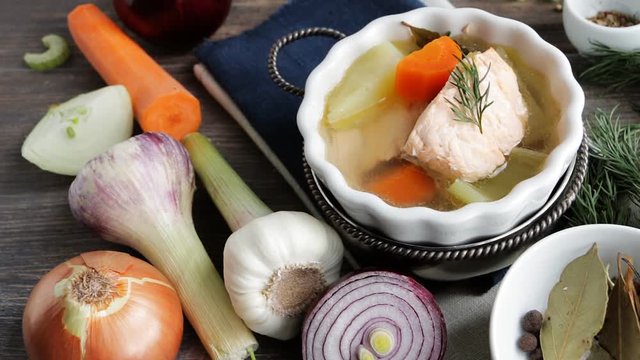Hot steaming fish soup on wooden table. Roots, vegetables, spices, condiments nearby. Slow pan.
