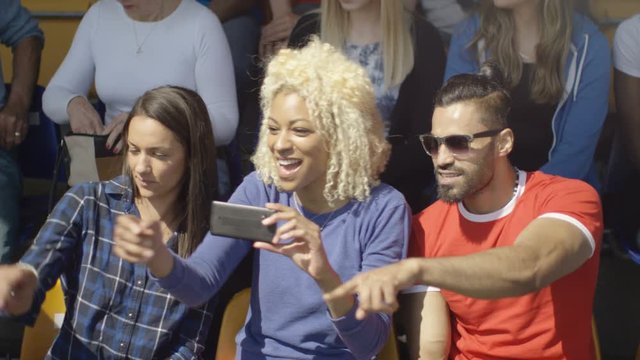  Friends sitting in the crowd at sports event taking photos with smartphone