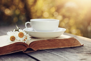 Old book and a cup with a camomiles on a wooden table.