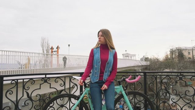 A young hipster girl standing with fixed gear bike at city centre near bridge, slow motion