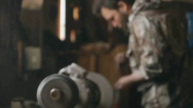 Dust fly in air on blurred background with bearded young man blacksmith manually polish metal knife on grinding machine in traditional smithy