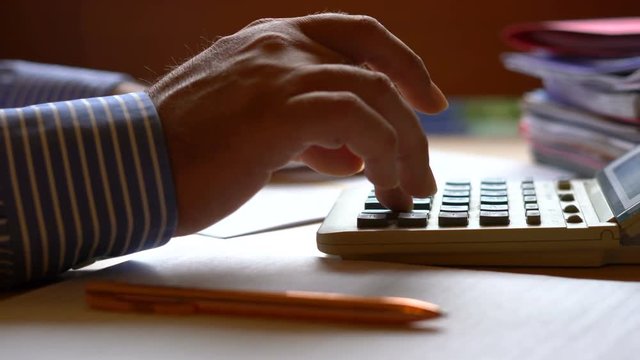 Accountant using calculator with stacks of paperwork and filing, in low light working long hours, macro closeup, dolly slider to calculator.