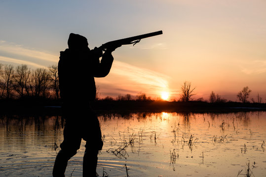 Silhouette of a hunter at sunset in the water with a gun.
