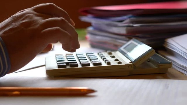 Accountant using calculator with stacks of paperwork and filing, in low light working long hours, macro closeup, dolly slider to hands.