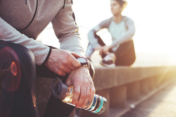 Two athletes resting after workout session on the street. Tired man with bottle tracking time with his arm watch and woman relaxing on the bench