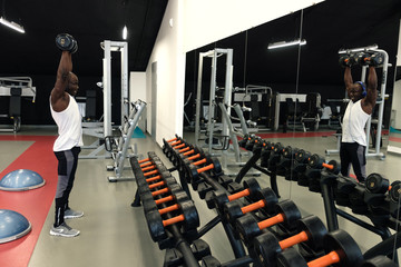 Muscular black man in a white t-shirt doing exercises with dumbbells in front of the mirror