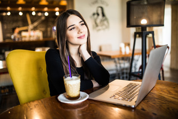 Young female with cute smile sitting with laptop in modern coffee shop interior during recreation time, charming happy woman student using laptop computer to prepare for the course work