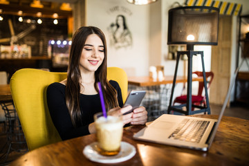 Young woman sitting in coffee shop at wooden table, drinking coffee and using smartphone. On table is laptop. Girl browsing internet, chatting, blogging.