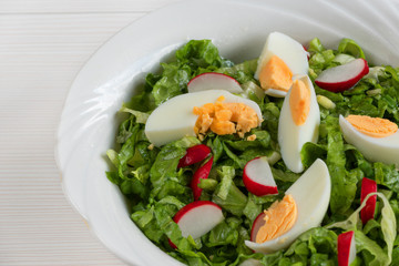 Close-up side view a colorful plate of green salad with lettuce, tomatoes, cucumber, onion, radishes and olives on white wooden board. Healthy food and eating concept. Fresh green salad background.