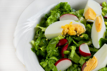 Close-up side view a colorful plate of green salad with lettuce, tomatoes, cucumber, onion, radishes and olives on white wooden board. Healthy food and eating concept. Fresh green salad background.