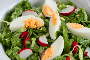 Close-up side view a colorful plate of green salad with lettuce, tomatoes, cucumber, onion, radishes and olives on white wooden board. Healthy food and eating concept. Fresh green salad background.