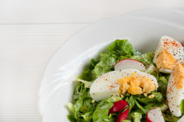 Close-up side view a colorful plate of green salad with lettuce, tomatoes, cucumber, onion, radishes and olives on white wooden board. Healthy food and eating concept. Fresh green salad background.