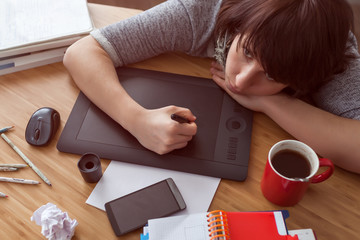 A girl draws a picture on a graphic tablet. She looks tiredly at the monitor. Various items on the table.