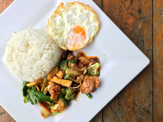 stir fried chicken with roasted chili paste and sweet basil leaf, Broccoli, mushroom mixed vegetables, Fried egg on jasmine rice in white plate on wooden background, thai food.