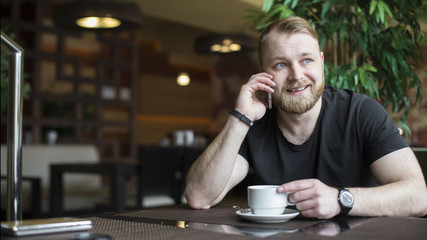 Young bearded attractive businessman in blue shirt is sitting at wooden table in restaurant, talking on phone and drinking tea. Man is waiting for colleague in cafe. Blurred background. Lifestyle.