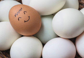 Closeup brown chicken egg with paint in smile face on pile of white duck egg on wood basket...