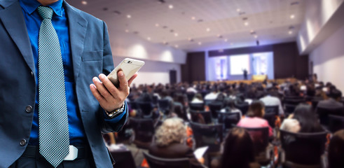 businessman using the mobile phone, blurred of conference hall or seminar room with attendee background.