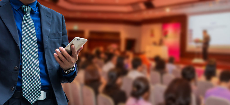 businessman using the mobile phone, blurred of conference hall or seminar room with attendee background.