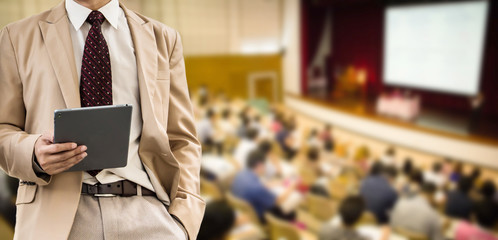 businessman using the tablet, blurred of conference hall or seminar room with attendee background.