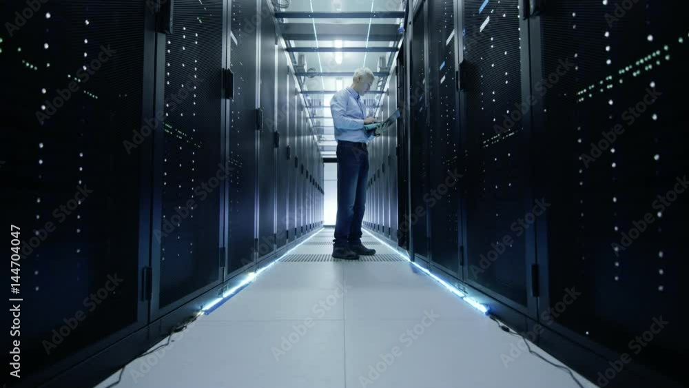 Poster data center door opens and we see server engineer working on a laptop. rack server cabinet is open. 