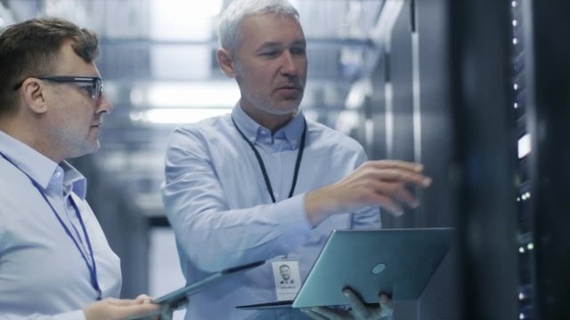  Two IT Technicians Standing in Working Data Center. They Use Laptop and Tablet Computer while Standing Beside Open Server Rack Cabinet. Shot on RED EPIC-W 8K Helium Cinema Camera.