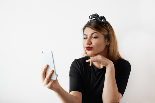 Portrait of pretty female student having fun and grimacing while taking selfie using mobile phone, posing on white wall background. Funny young woman makes a duck face posing indoor. Selective focus