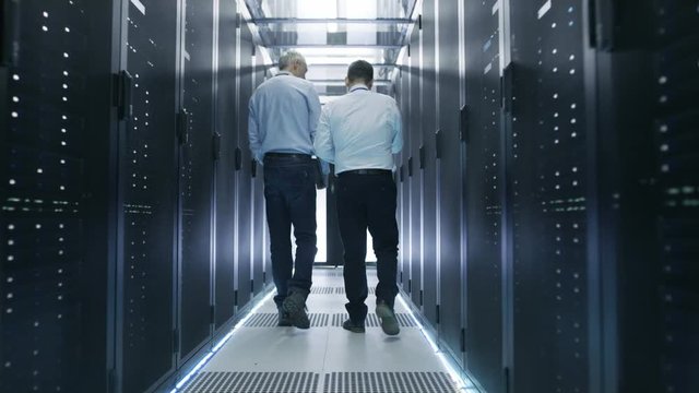Two Server Technician Working in Data Center. One Uses Tablet Computer. They Walk Through Rows of Server Racks. Shot on RED EPIC-W 8K Helium Cinema Camera.