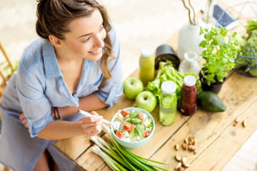 Young and happy woman eating healthy salad sitting on the table with green fresh ingredients indoors