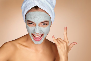 Funny woman wearing a facial mask and making rock hand gesture. Portrait of young woman on on beige background. Skin care concept