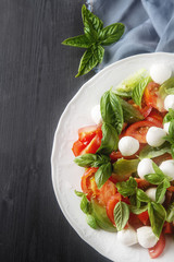 White plate with a light summer salad of tomato, mozzarella, basil, spices, olive oil. Dark wood background. Blue cloth. free space text. Top view.