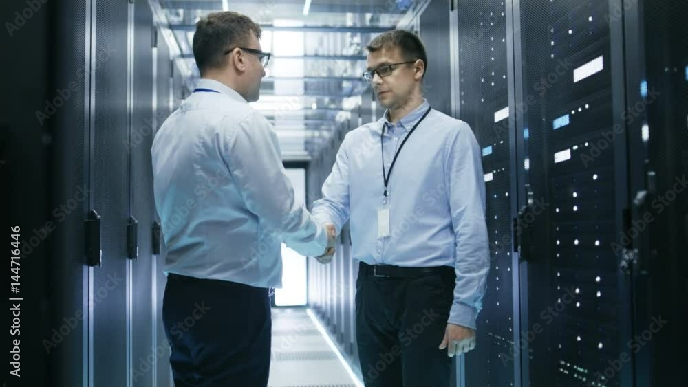 Poster two corporate workers shake hands in greeting. they're working in modern data center with rows of se