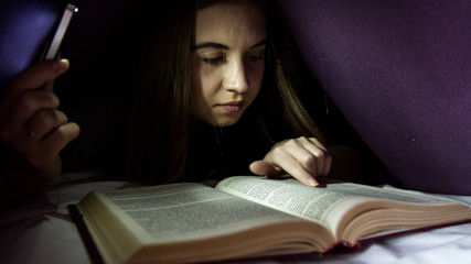 Young woman hiding under blanket and enrapt reading interesting book at nighttime. Girl lighting...
