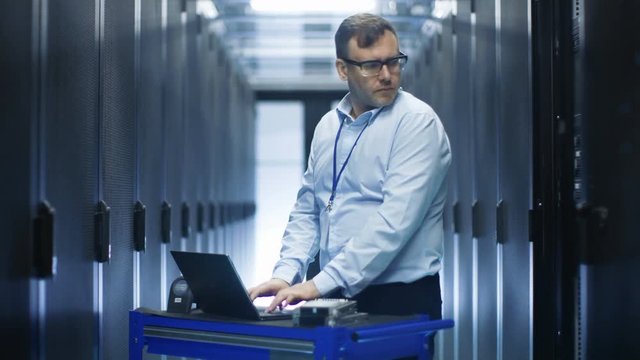 In Data Center IT Engineer Works on Crash Cart Laptop. We See Rows of Server Racks and One Cabinet is Open. Shot on RED EPIC-W 8K Helium Cinema Camera.