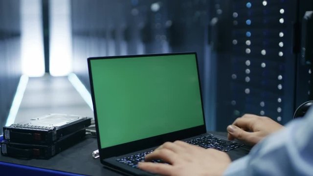 IT Technician is Working on a Green Screen Chroma Key Laptop in Big Data Center with Rows of Server Racks in It.Shot on RED EPIC-W 8K Helium Cinema Camera.