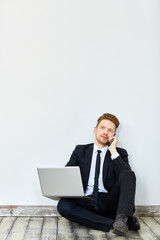 Portrait of handsome young red haired businessman using laptop and speaking by phone sitting on floor of empty office room