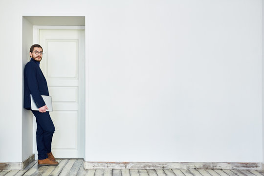 Bearded Man In Glasses Leaning On Doorway Holding Laptop Computer And Looking At Camera Next To Blank White Wall In Office