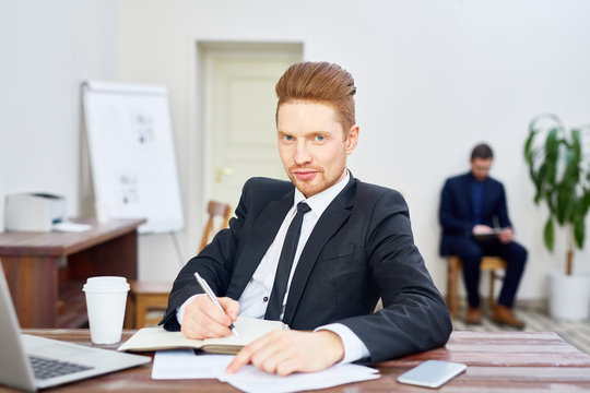 Portrait Of Handsome Young Businessman Looking At Camera With Smirk While Writing In Planner At Desk In Office, Employee In Background