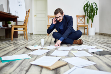 Portrait of middle aged bearded man speaking by phone and sorting documents sitting on floor in...