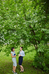 Portrait of two young women dressed in business clothes chatting under big tree in nature during break
