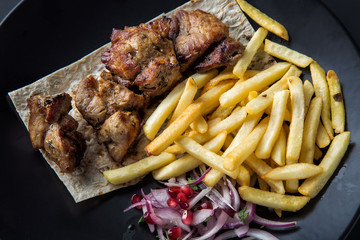 Grilled shish kebab served with french fries, onion and pomegranate grains on black plate close up